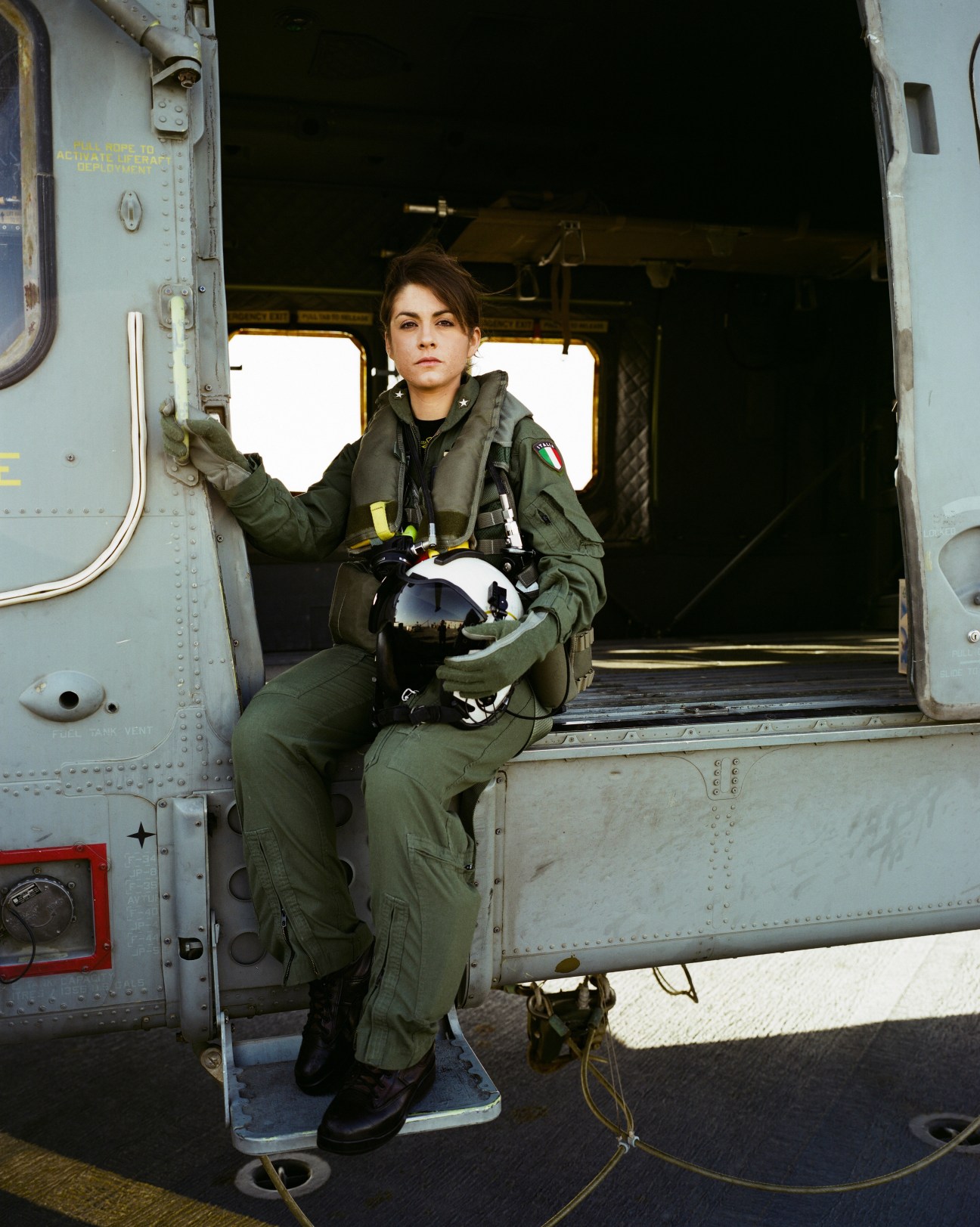 Helicopter pilot Carmela Carbonara in flight suit sitting in helicopter doorway holding helmet.