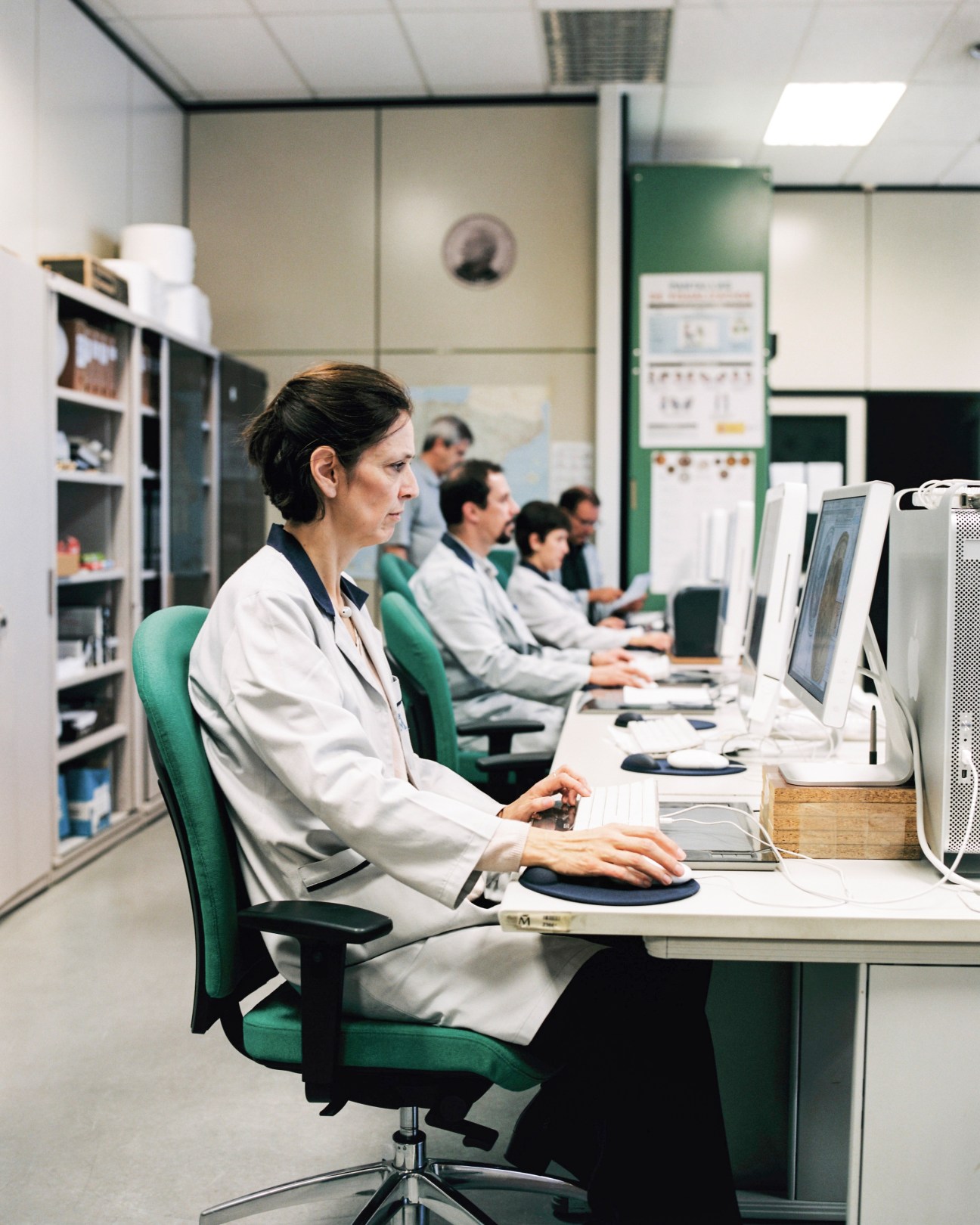 Woman in white lab coat at computer workstation in mint facility with colleagues at desks behind her.