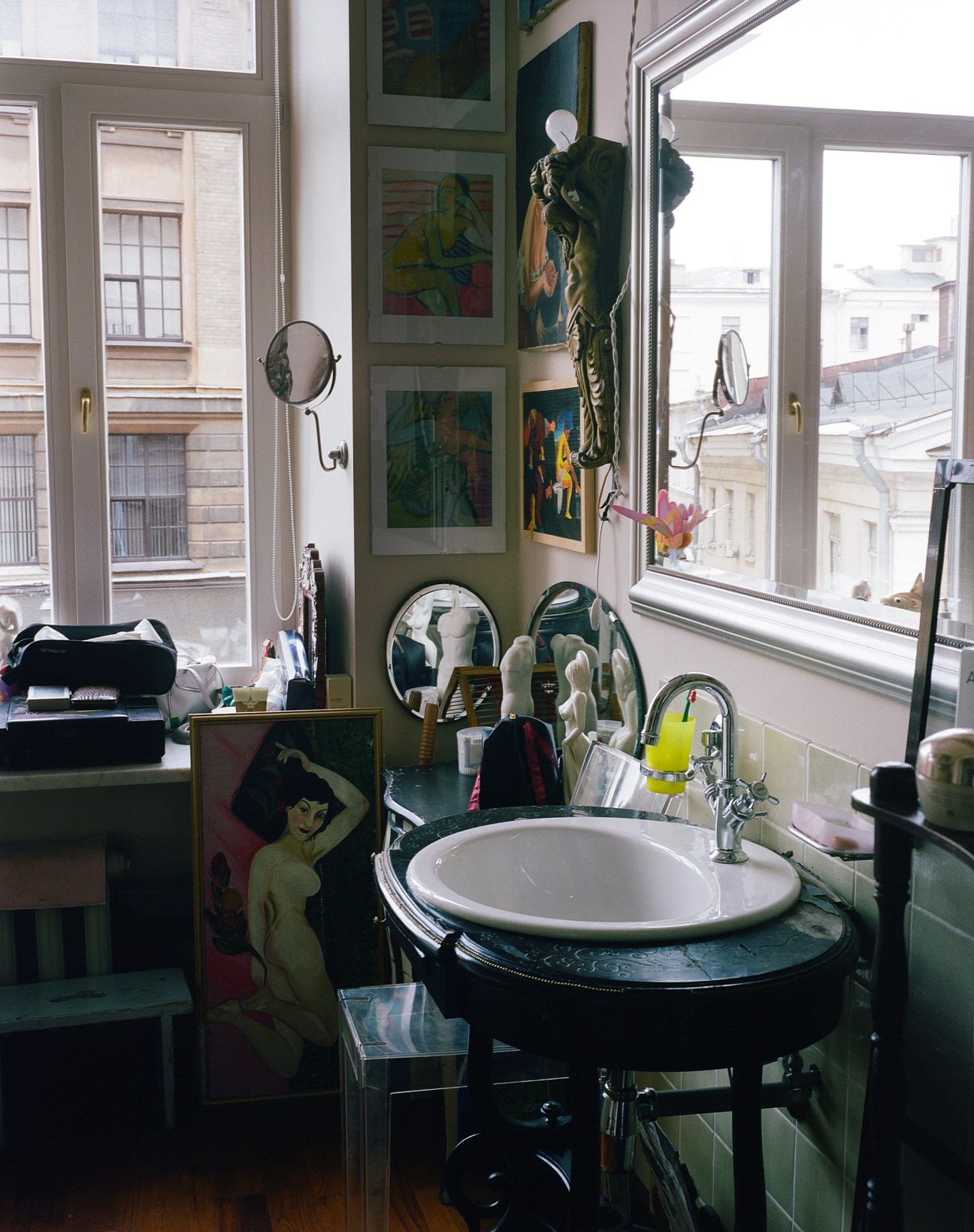 A bathroom with green walls, white sink, multiple mirrors, artwork, and large windows overlooking St Petersburg buildings.