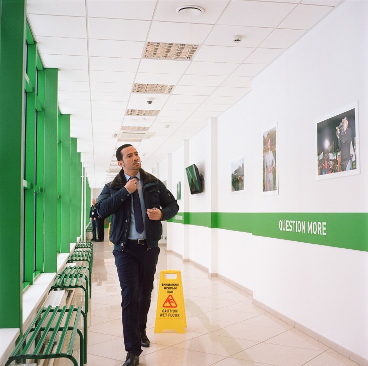 Man in dark coat walking through corridor with green benches and 'Question More' branding on wall.