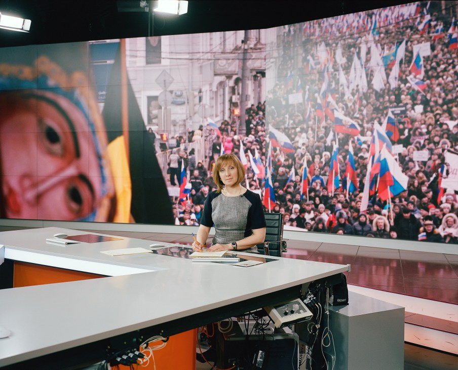 News anchor Marina Dzhashi sits at RT studio desk with large screen behind showing Russian crowd with flags.
