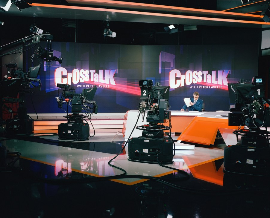 RT's main studio in Moscow with presenter at desk, multiple cameras, and orange and black set design with 'CrossTalk' branding visible on screens.