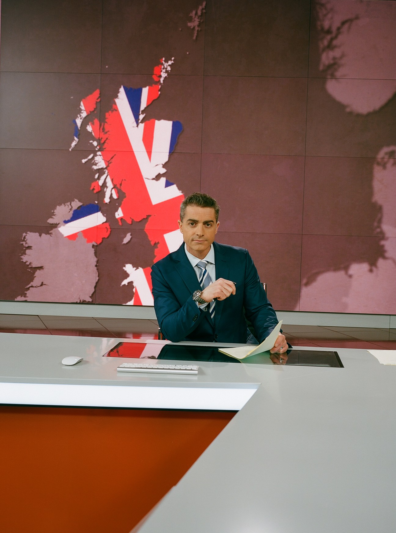British news anchor Rory Suchet sits at an RT studio desk with a UK map displayed behind him.