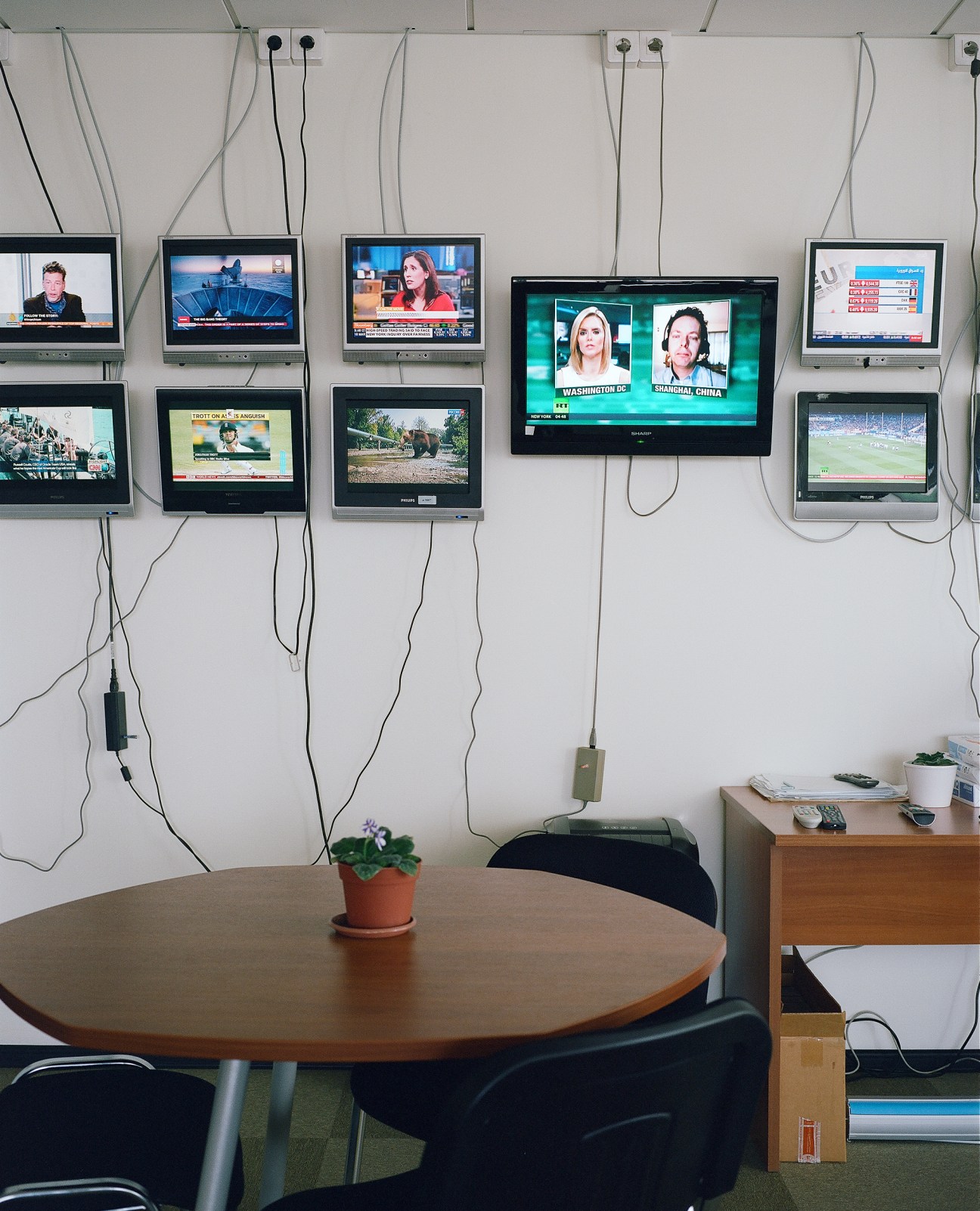 Multiple television monitors mounted on a white wall showing various news broadcasts and feeds, above a wooden table in an RT control room.