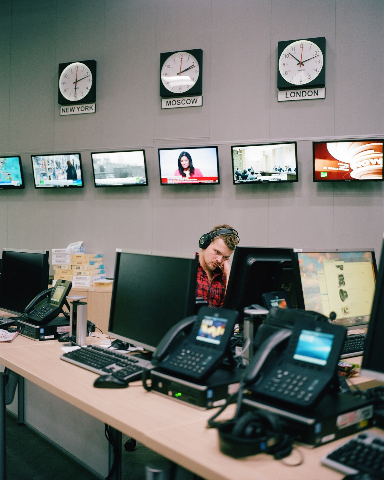 Editor at desk in RT studio with multiple monitors, phones, and wall clocks showing New York, Moscow and London times.
