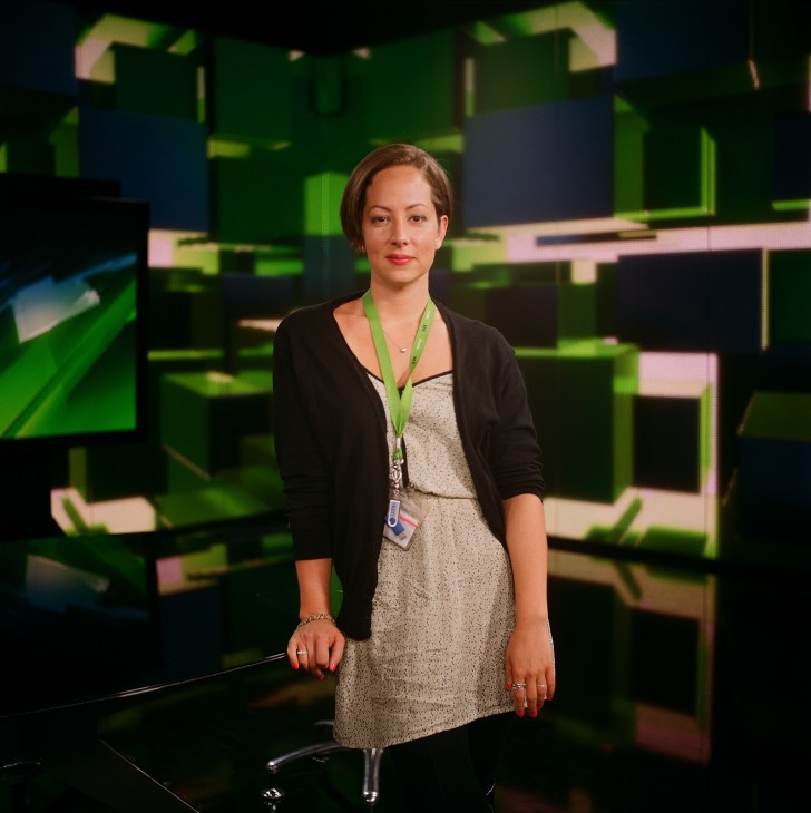 American presenter Anissa Naouai stands in RT's studio with green and blue set elements behind her.