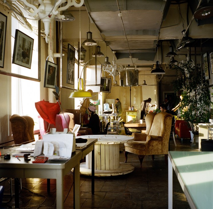 Interior of Etagi art space showing industrial ceiling with pendant lights, vintage furniture, framed artwork, and visitors browsing.