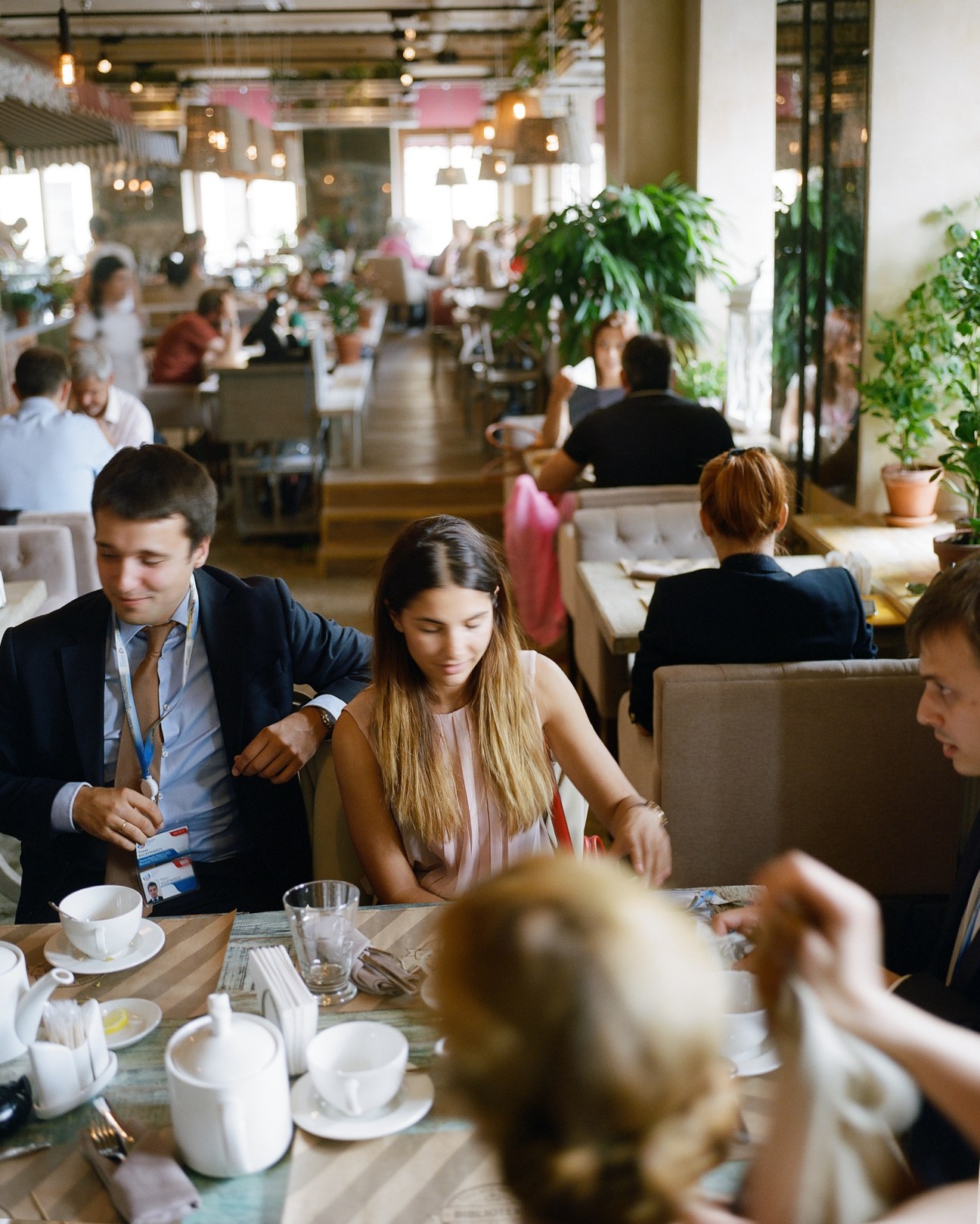 People dining in a bright, spacious restaurant with hanging lights and green plants.