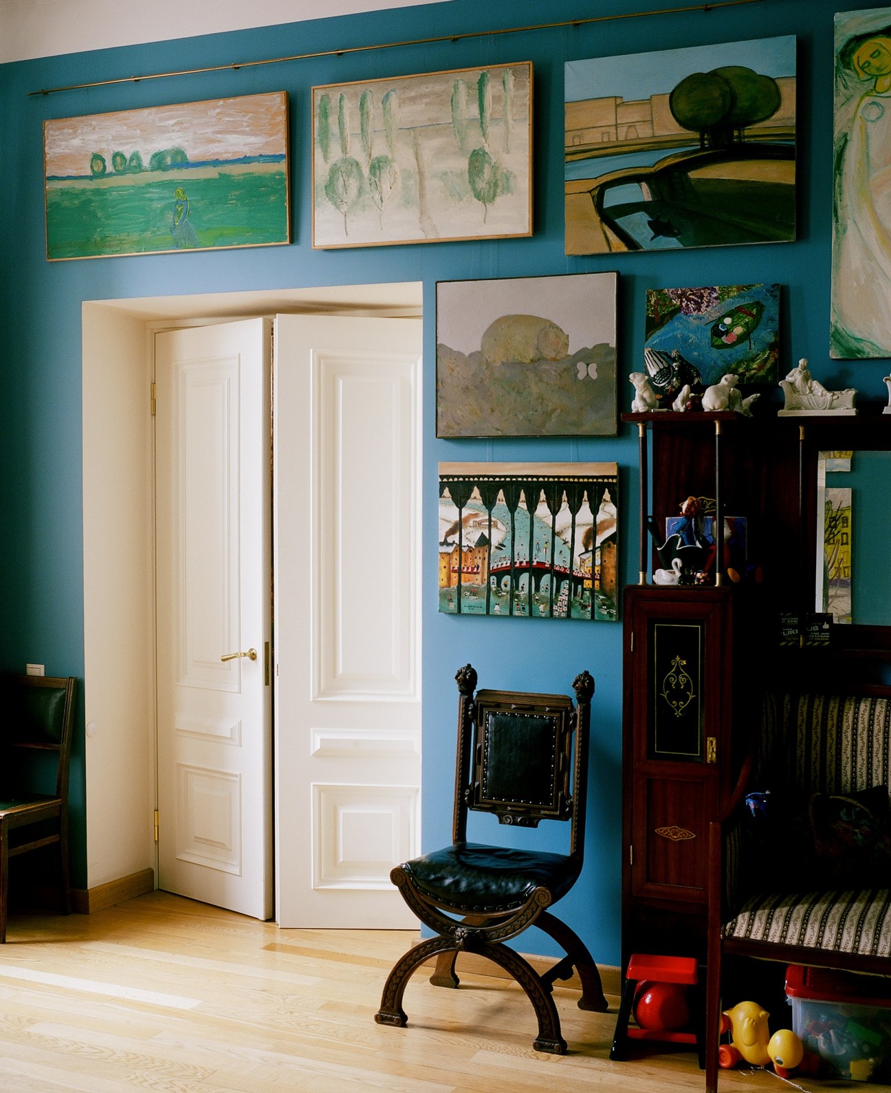 Colorful artworks hung on a blue-green wall above antique furniture and decorative objects in a St Petersburg apartment.