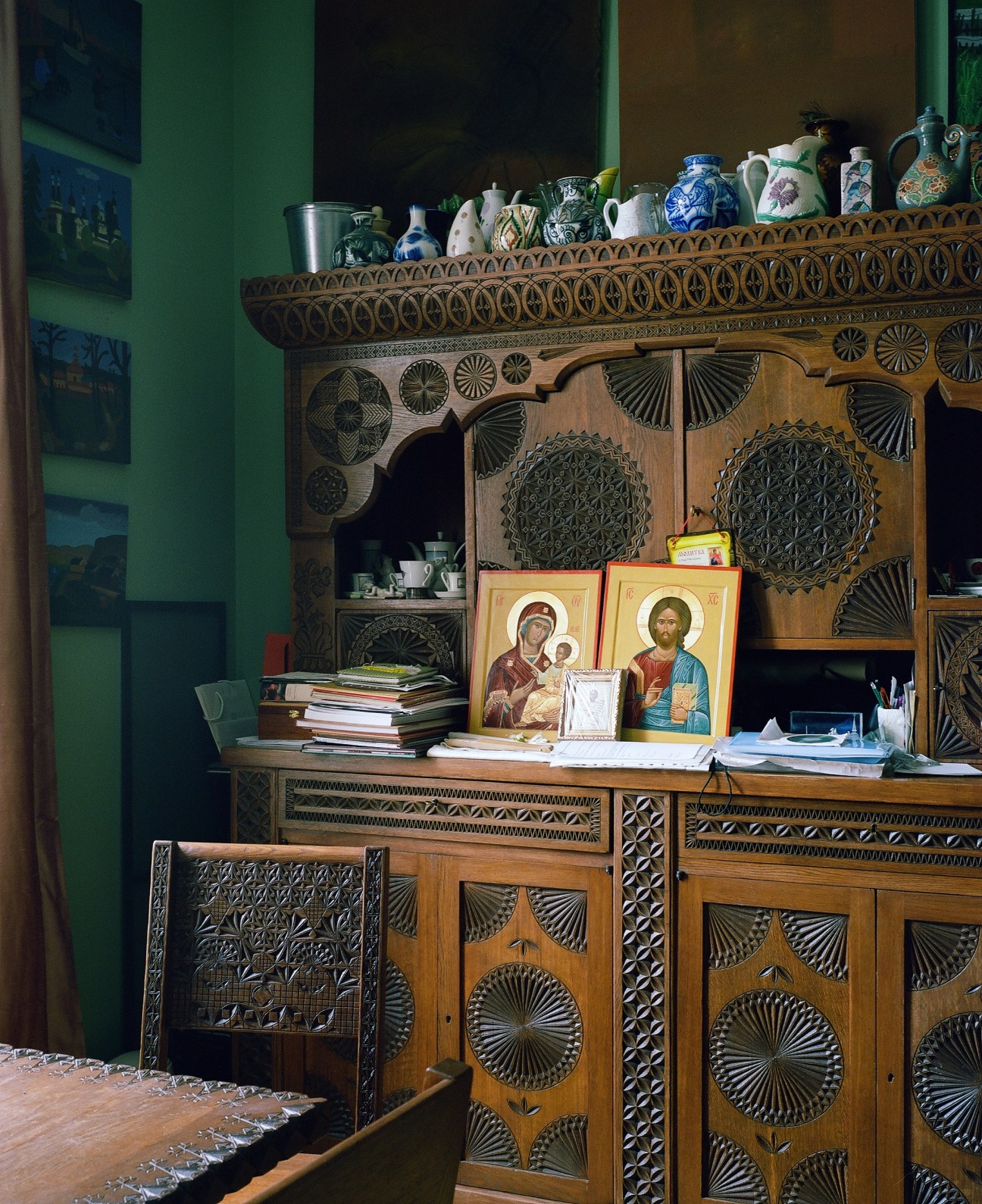 Ornate wooden cabinet with carved decorative panels displaying porcelain vessels and religious icons against a green wall.