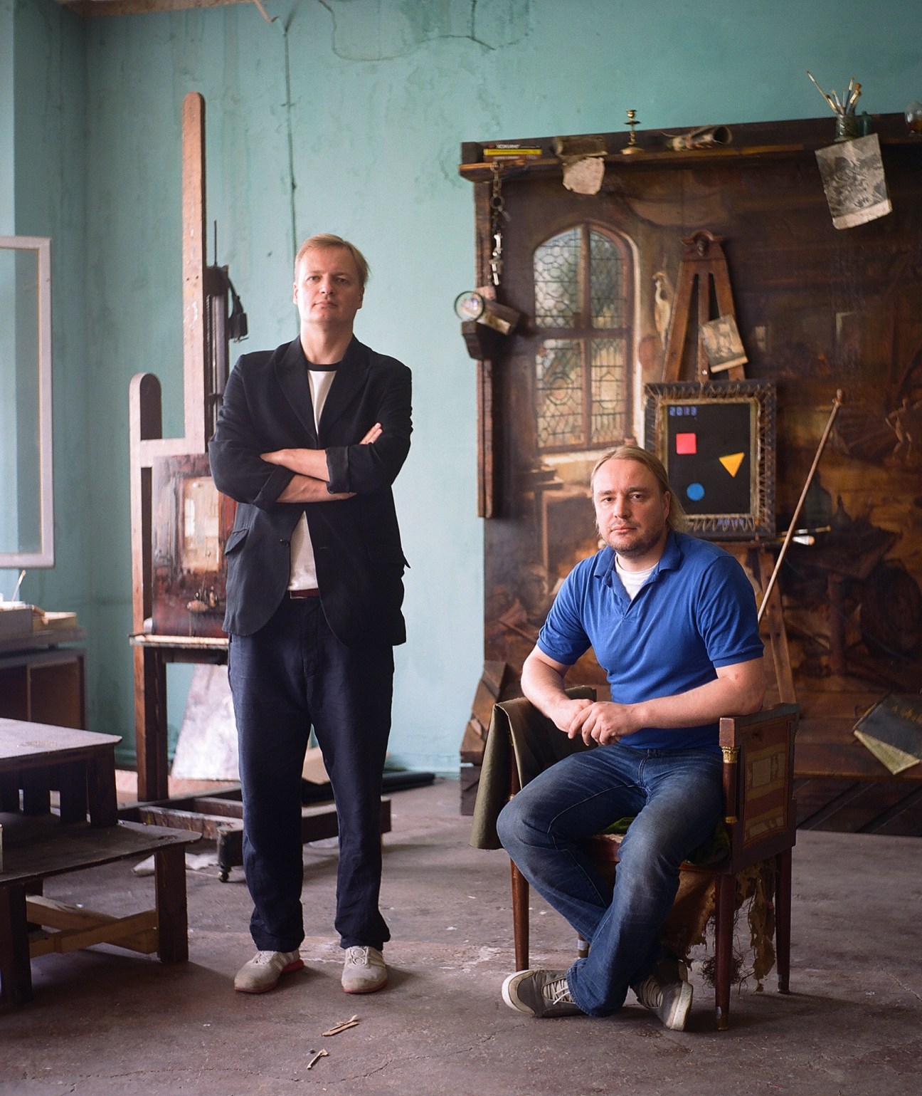 Architects Savely and Egor Archipenko pose in a studio space with turquoise walls and wooden furniture.