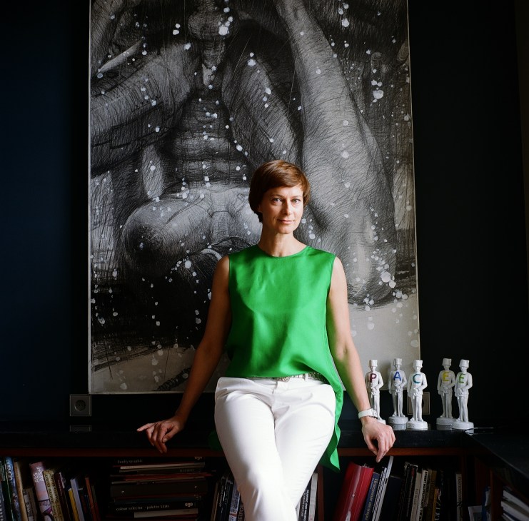 Marina Gisich, a woman with reddish hair wearing a green sleeveless top and white pants, sits on a shelf in front of abstract artwork.