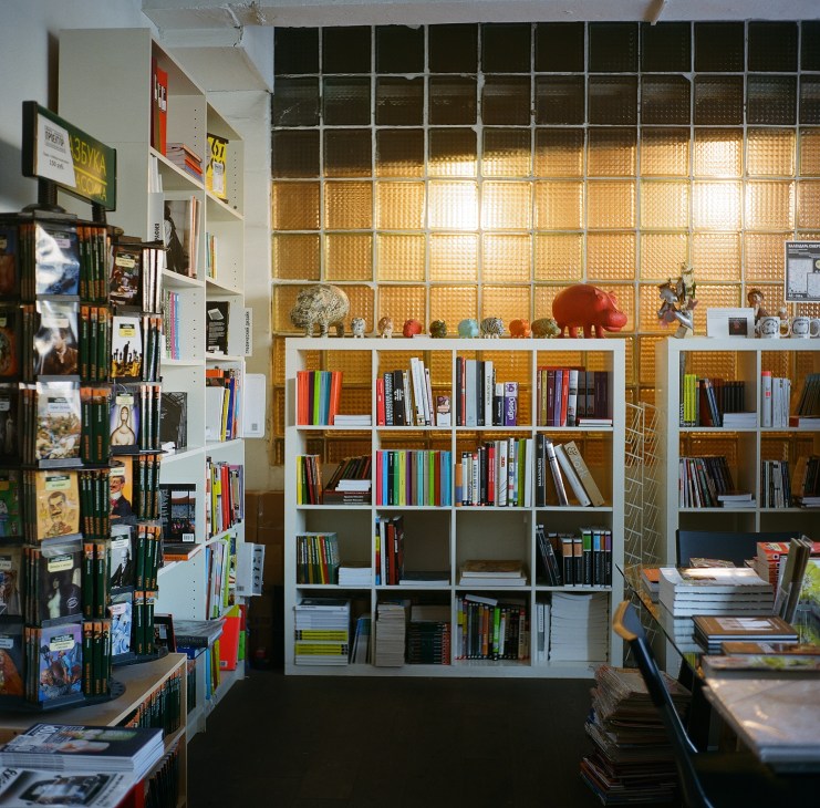 Interior of a bookshop with white shelving units filled with colorful books and decorative objects against a textured golden wall.