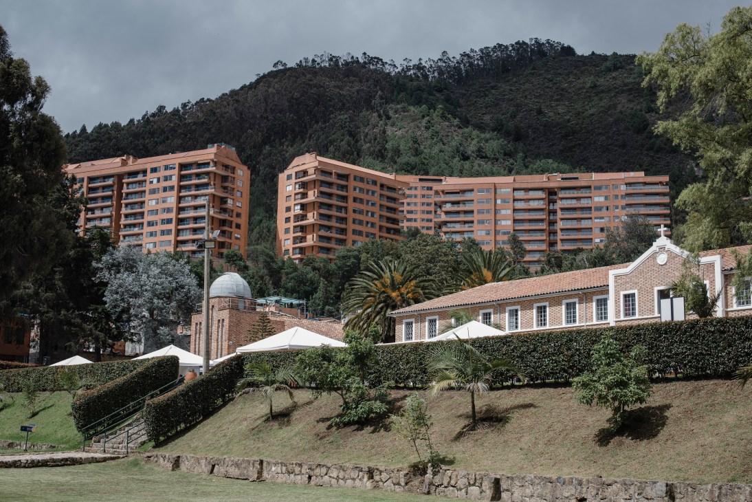 Brick monastery with white trim and modern apartment buildings rising behind it in a forested hillside setting.