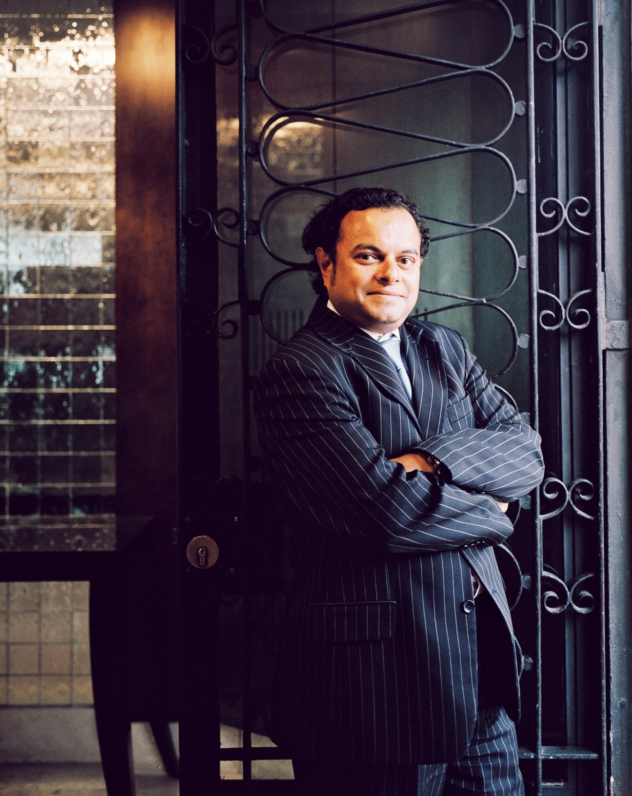 Man in pinstriped suit standing with arms crossed against ornate metal door.