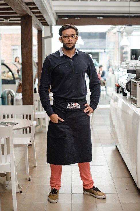 Man in dark polo shirt and black apron standing in modern café interior