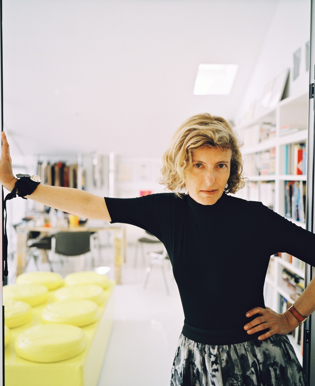 Teresa Sapey, Italian architect, posing in her bright modern office with bookshelves and yellow furniture.