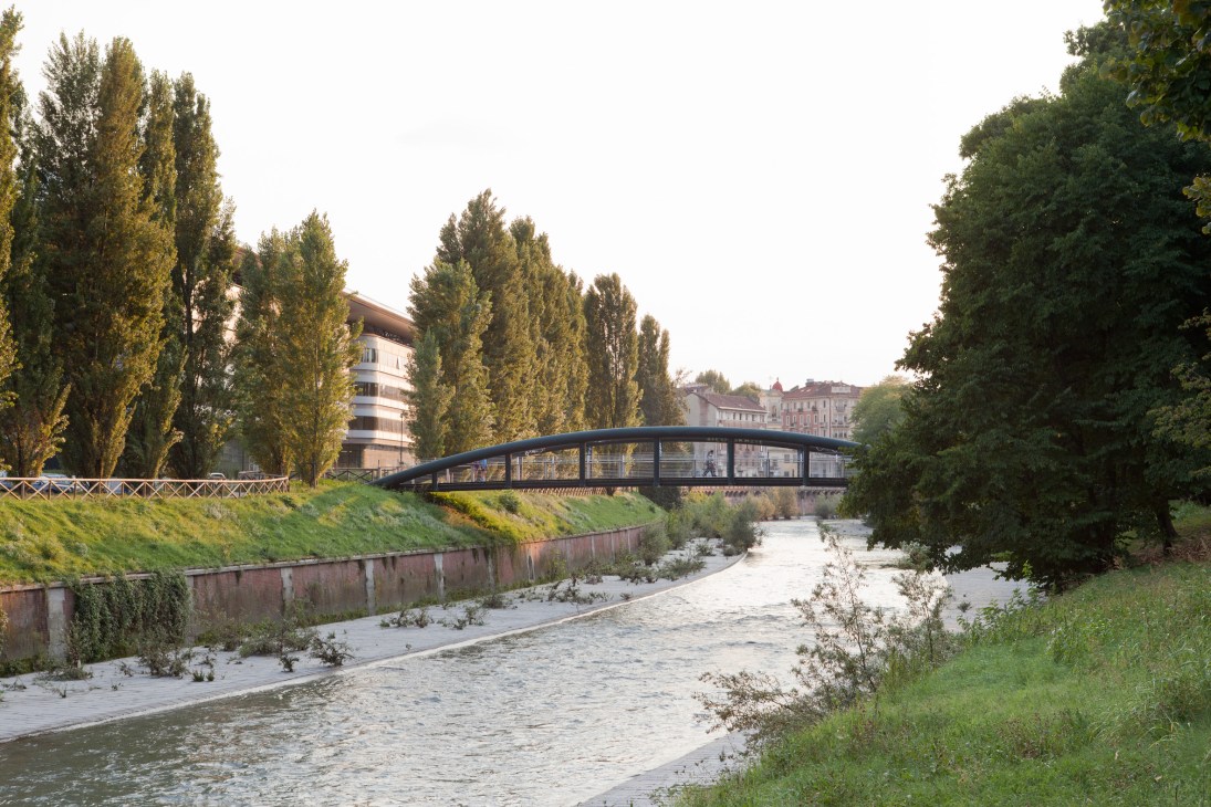 Modern curved pedestrian bridge spanning the Dora Riparia River lined with tall poplar trees in Turin.