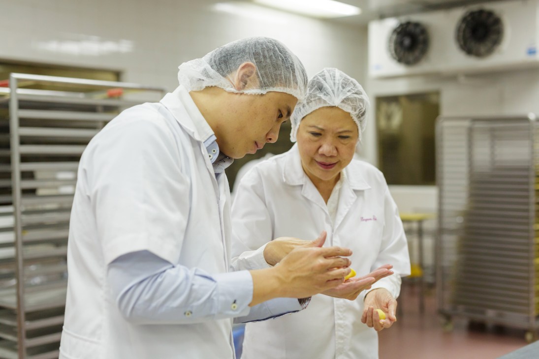 A man and woman in white chef coats and hair nets examine a pastry in a commercial kitchen.