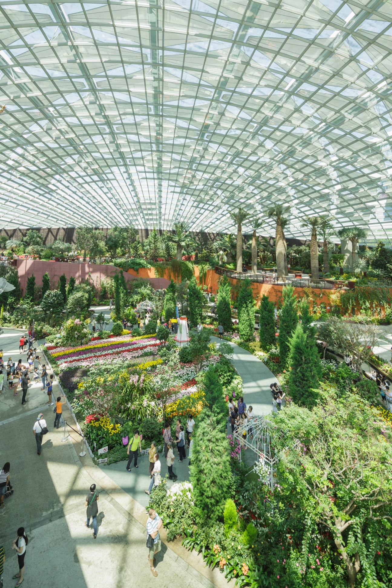 Interior view of Gardens by the Bay's Flower Dome biome with glass ceiling, colorful flower beds, and visitors walking through lush tropical plants.