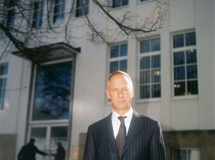 Wilhelm Schmid, CEO of A Lange & Söhne, stands in a pinstriped suit outside a modernist building with bare branches overhead.