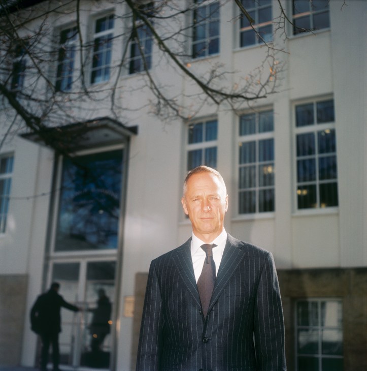 Wilhelm Schmid, CEO of A Lange & Söhne, stands in a pinstriped suit outside a modernist building with bare branches overhead.