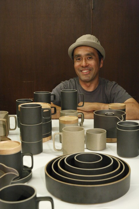 Designer Taku Shinomoto smiles behind a display of Hasami porcelain tableware featuring black and natural ceramic vessels with wooden accents.