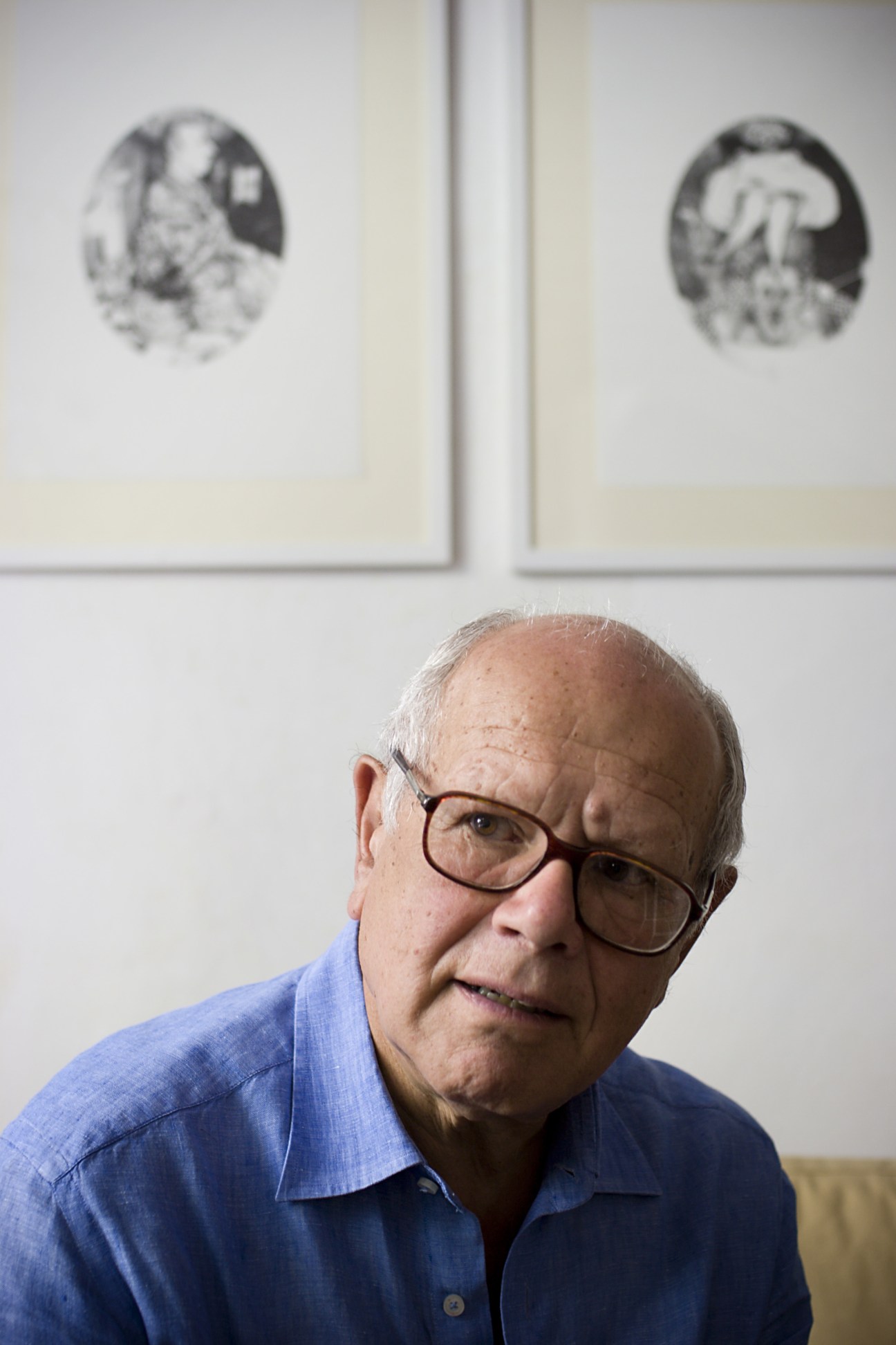 Portrait of Emilio Giannelli, an elderly man wearing glasses and a blue shirt, with his cartoon sketches visible on the wall behind him.