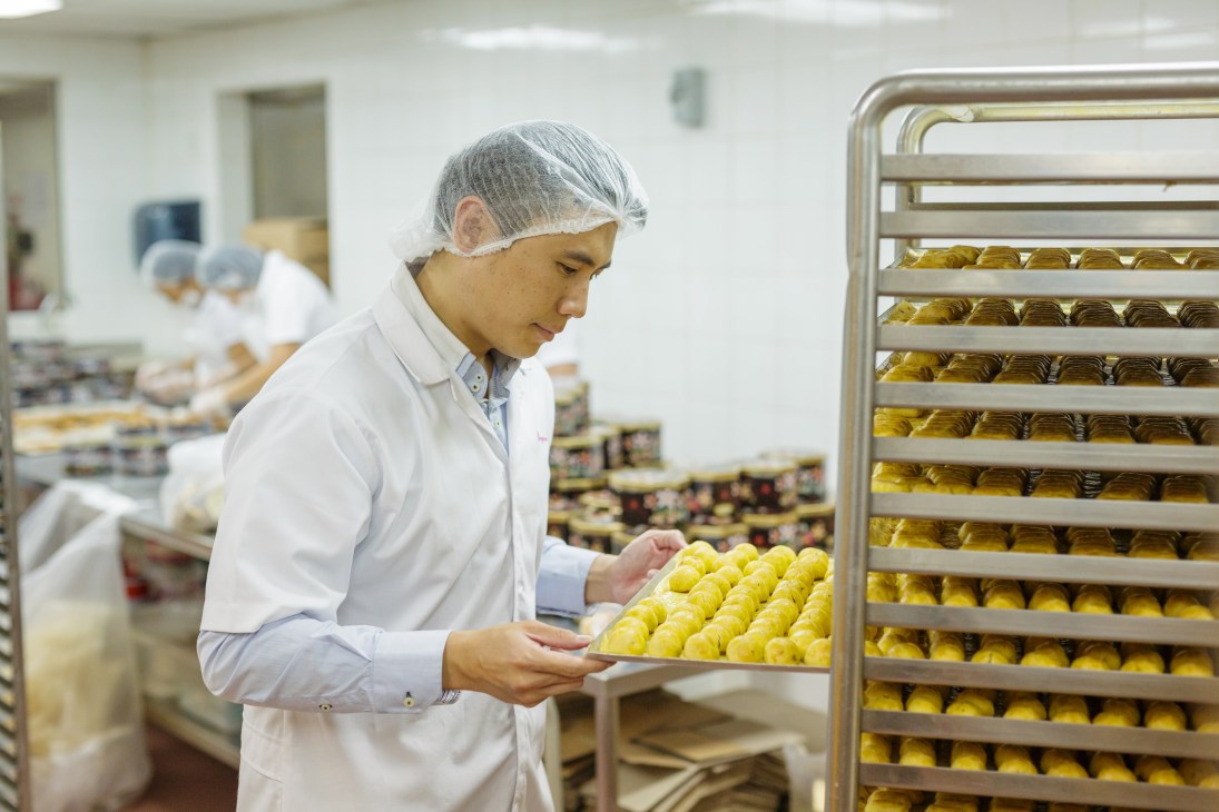 A baker in white coat and hair net inspects yellow pineapple tarts on a cooling rack in Bengawan Solo's kitchen.