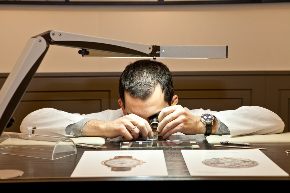 A Lange & Söhne watchmaker examining watch components at a workbench under an adjustable lamp.
