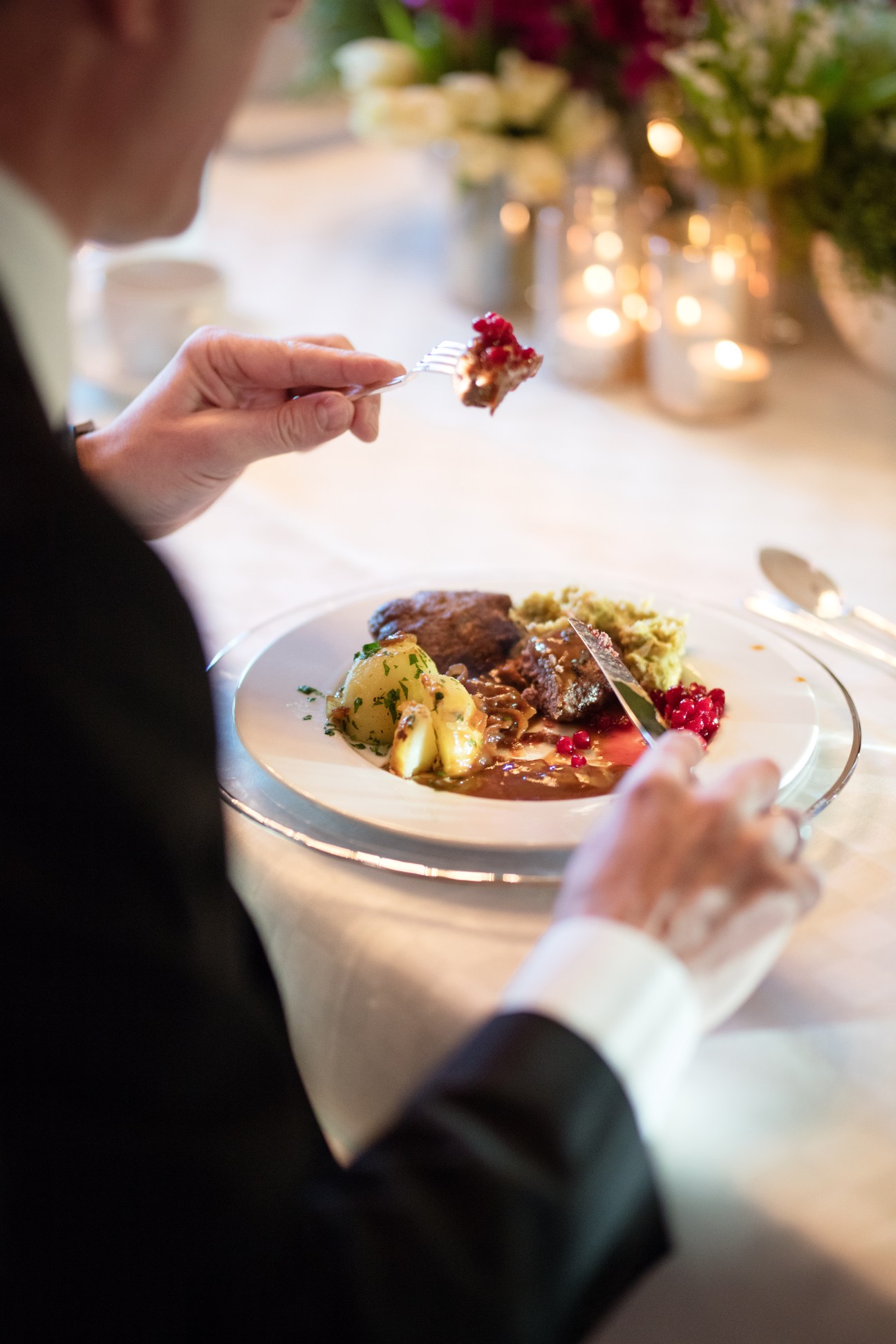 Person holding a fork with a bite of food above a white plate of fine dining meal.