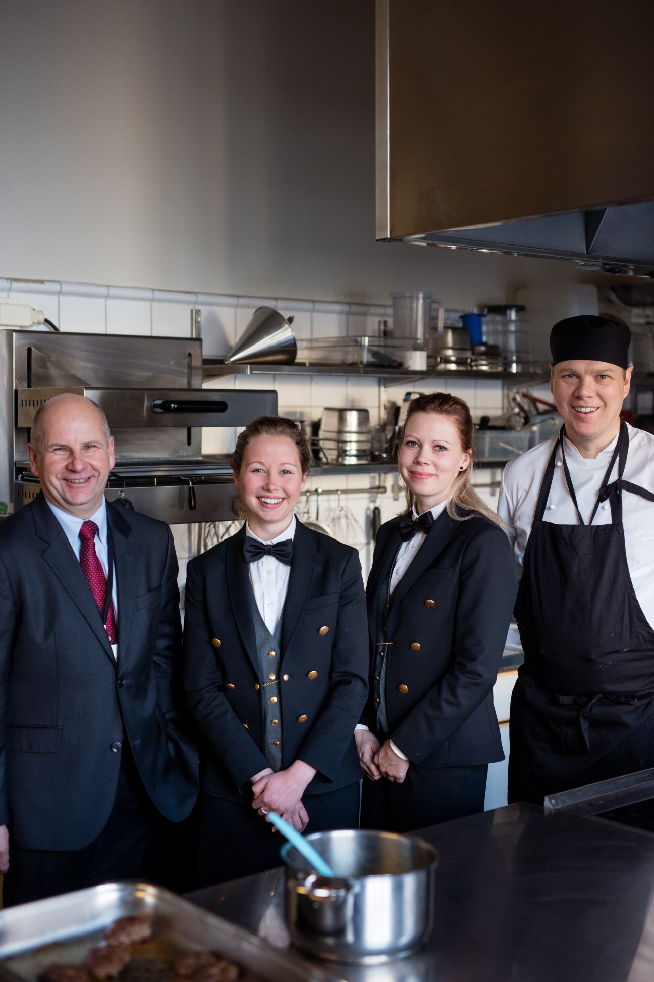 Four catering staff in formal attire stand in a professional kitchen with stainless steel equipment.