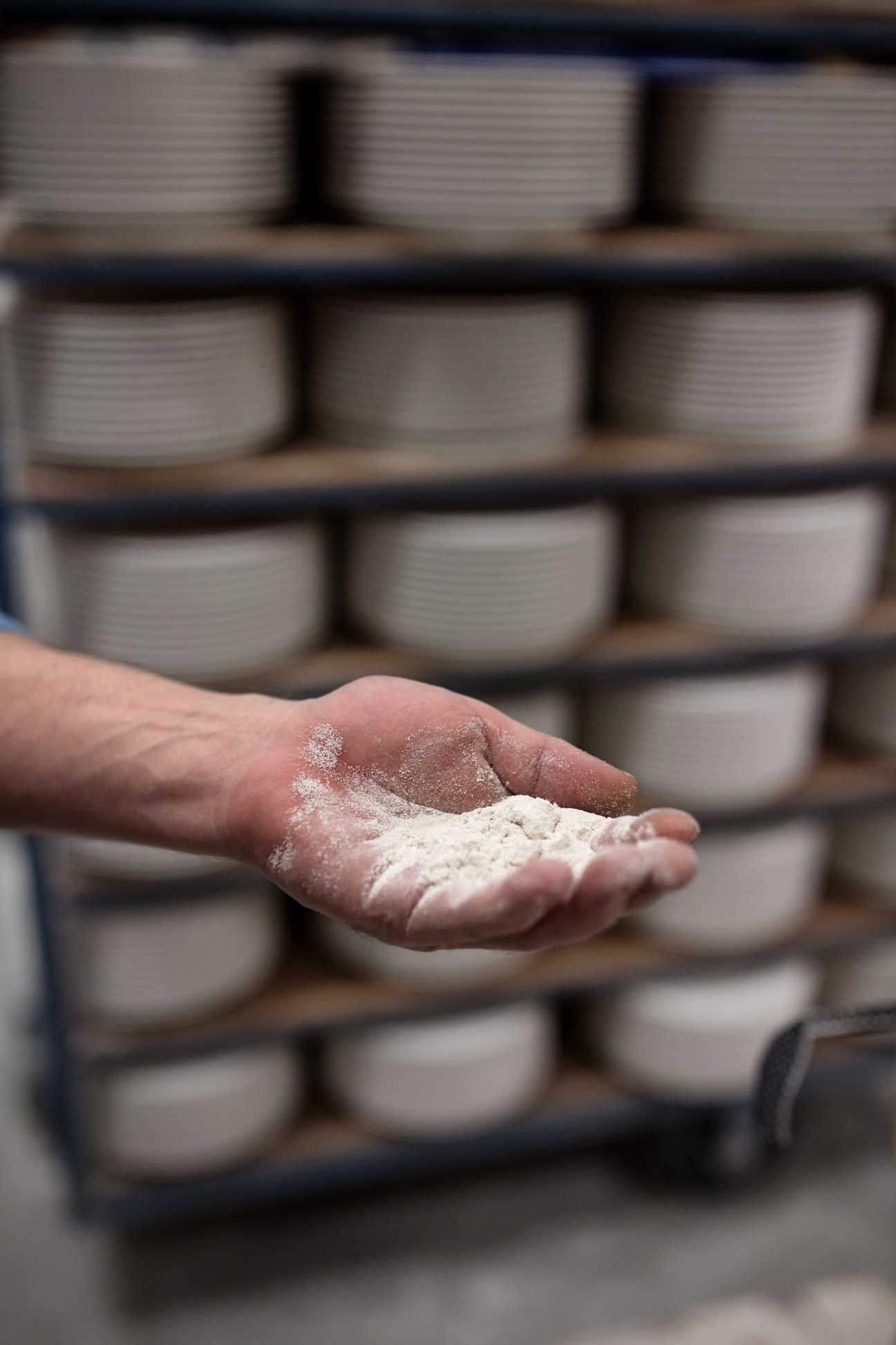 Hand holding raw porcelain clay powder with shelves of white ceramic bowls blurred in background