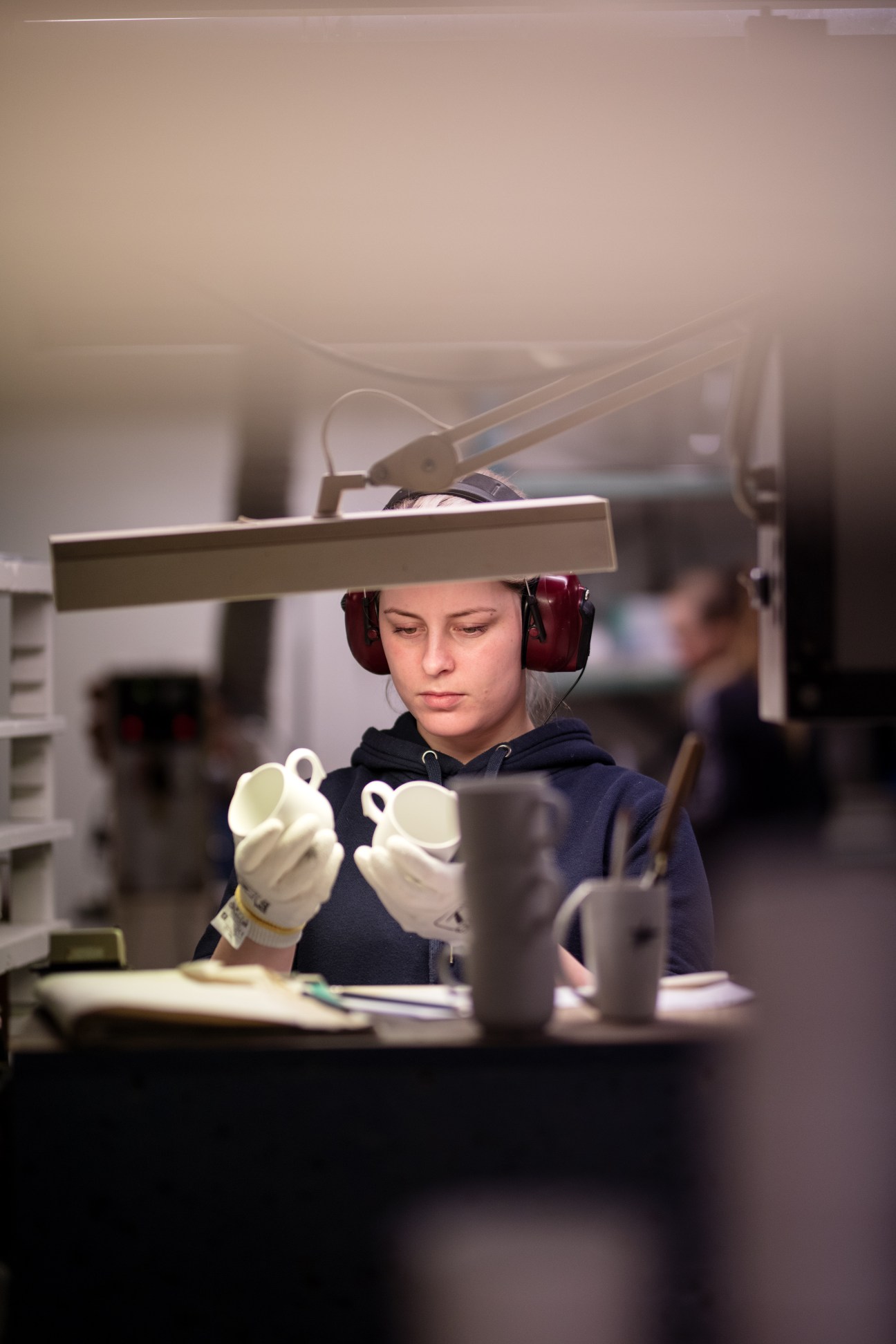 Worker at Figgjo factory wearing headphones and white gloves inspects white porcelain cups during quality control.