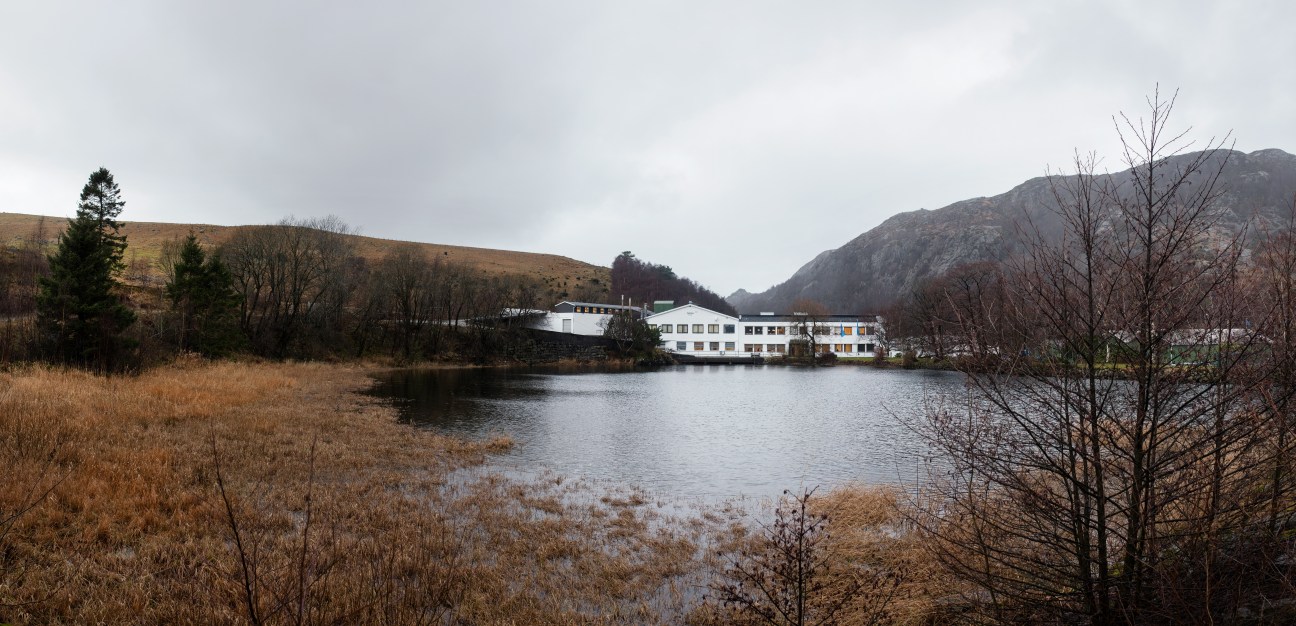 White modernist factory building on the shore of a calm lake surrounded by hills and bare trees in winter.