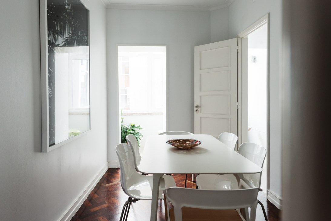 A bright, minimalist dining space with white walls, a white table, modern chairs, and natural light from doors and windows.