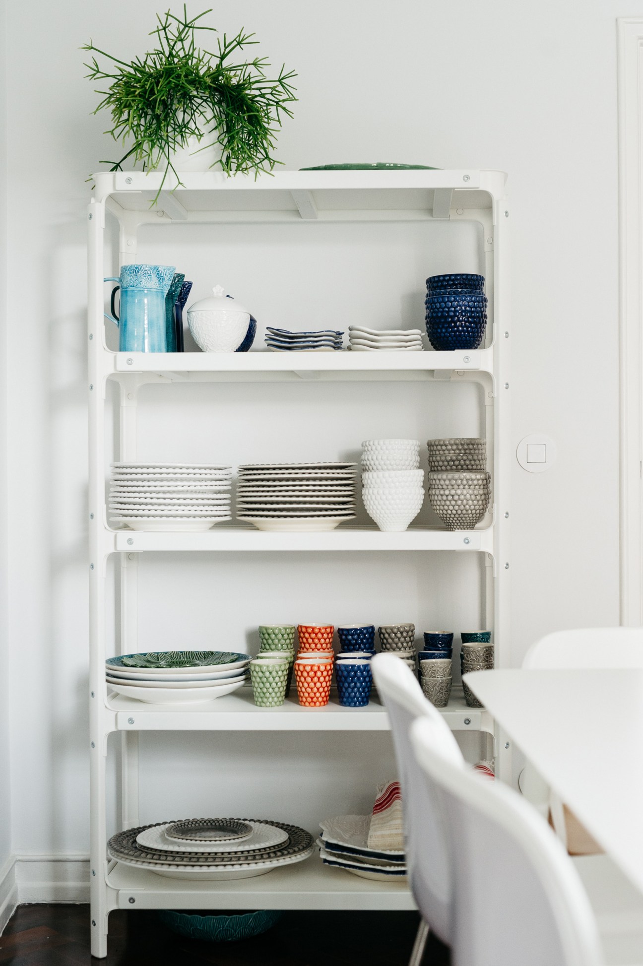 White wall-mounted shelving unit displaying organized dinnerware, glassware, and decorative items with a hanging plant on top.