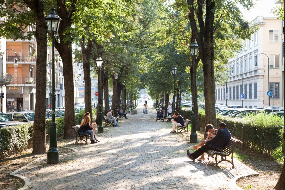 Tree-lined pedestrian walkway with benches, lamp posts, and people sitting under dappled sunlight in an urban square.