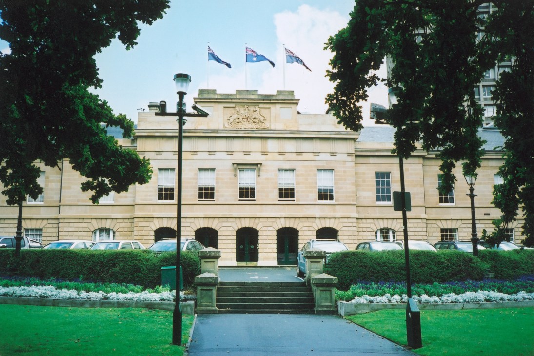 Parliament House in Hobart with three flags flying on the roof, framed by ivy-covered trees and manicured gardens.
