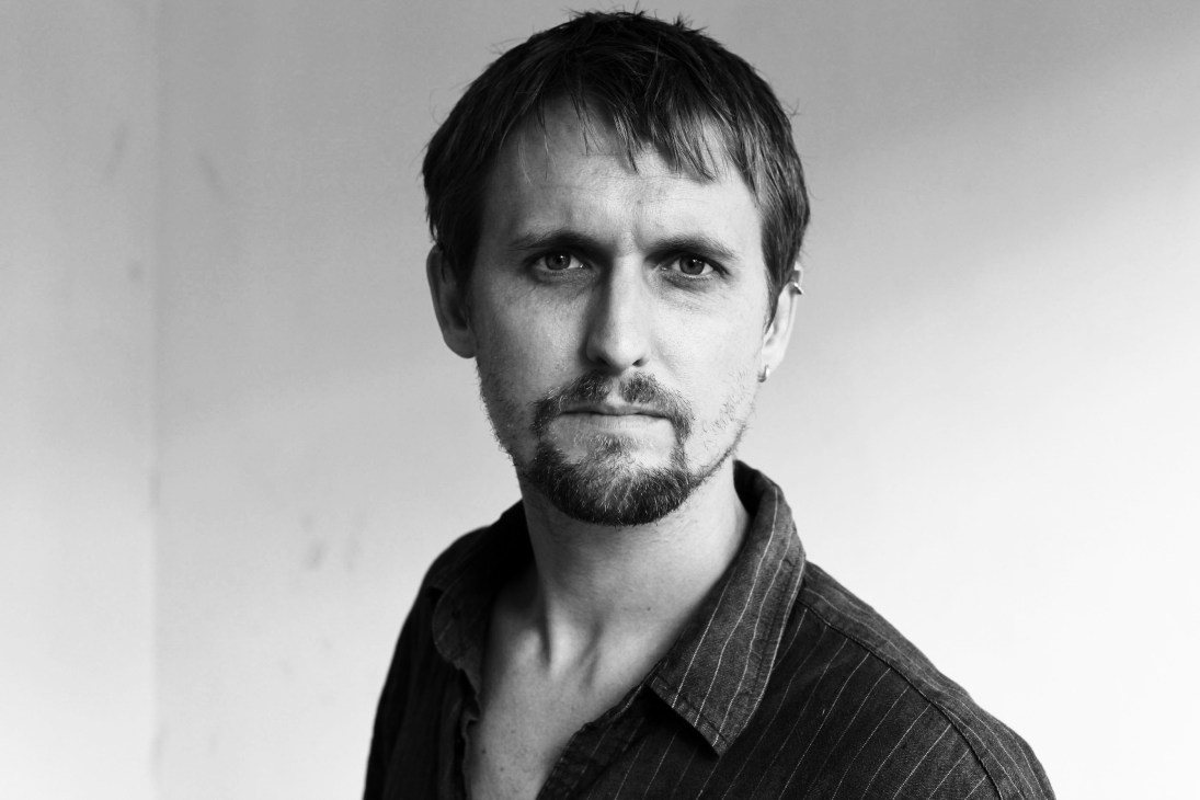 Black and white portrait photograph of a man with dark hair and facial hair wearing a dark pinstriped shirt.