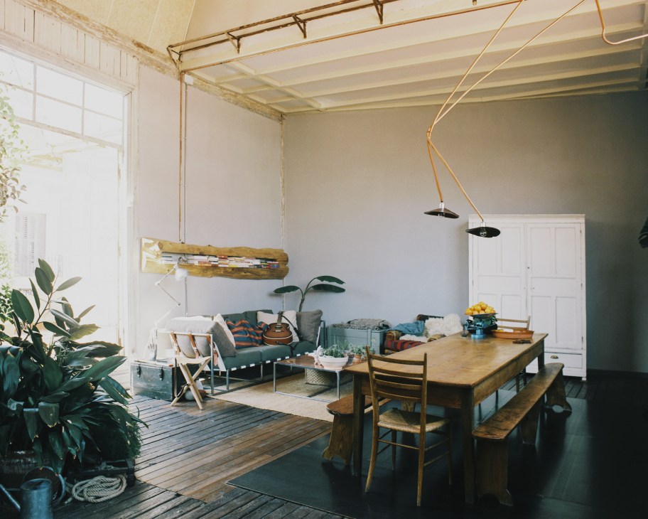 Open-plan living and working space with wooden dining table, green sofa, potted plants, and industrial brass light fixtures.