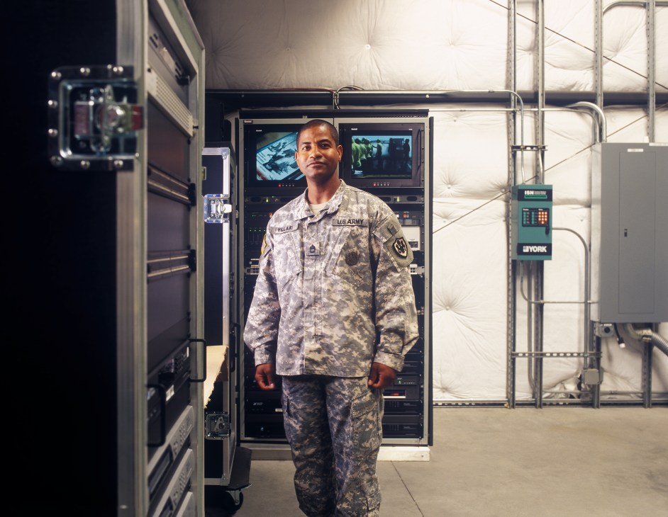 Sergeant Antonio Villari in U.S. Army camouflage uniform stands in front of deployable broadcast equipment and servers.