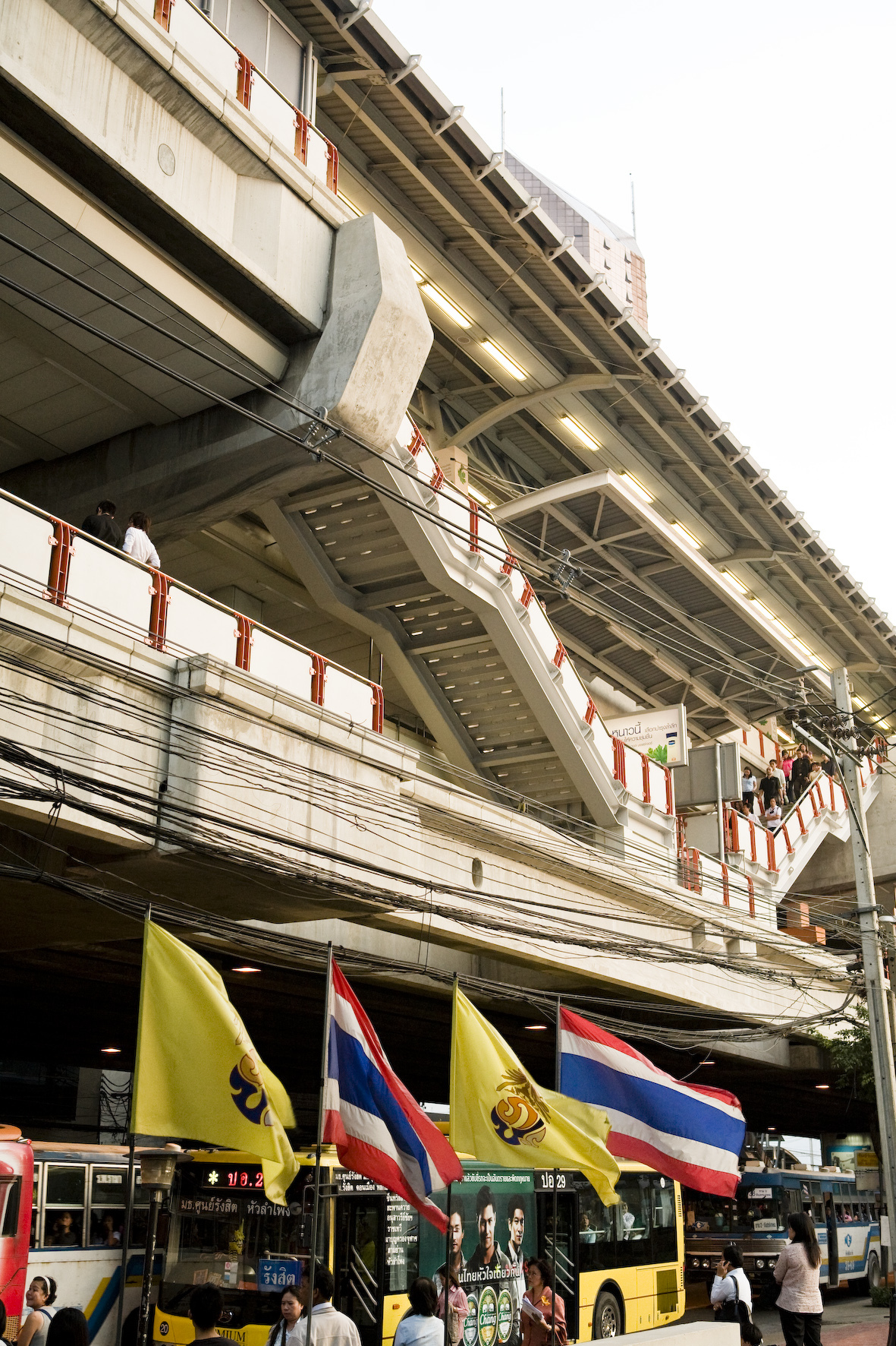 Bangkok's BTS SkyTrain elevated station with concrete structure and red railings above street-level shops and yellow buses.