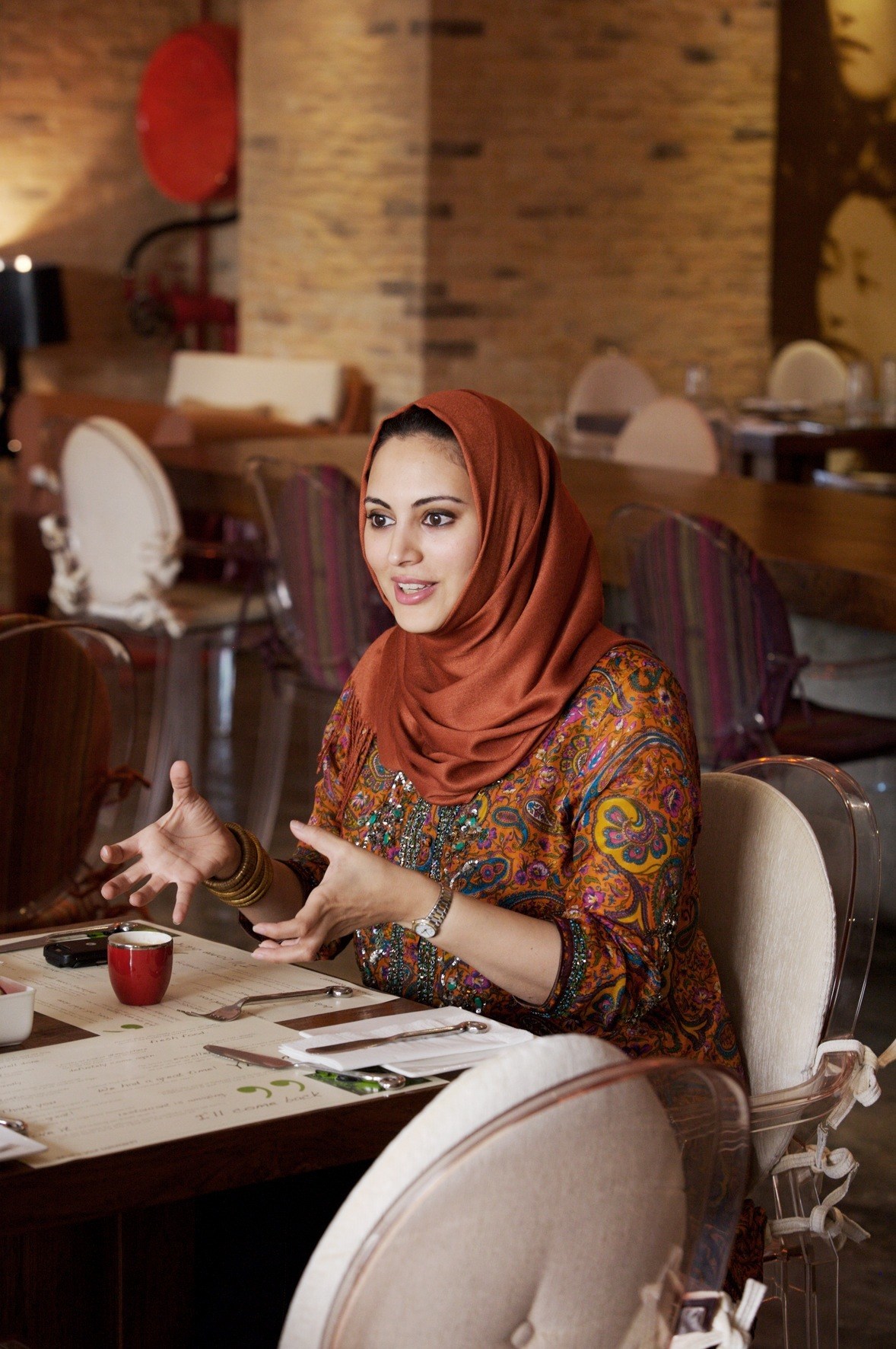 Muna Abu Sulayman seated at a restaurant table, wearing a rust-colored hijab and patterned top, gesturing while speaking.