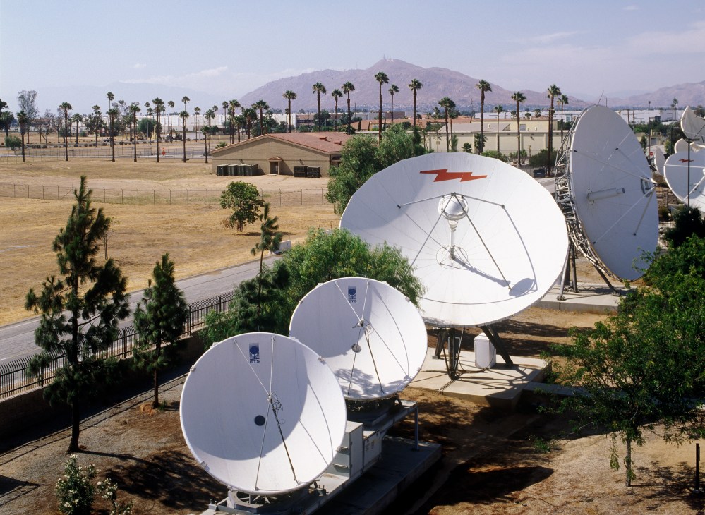 Satellite dishes in a desert facility with palm trees and mountains in the background.