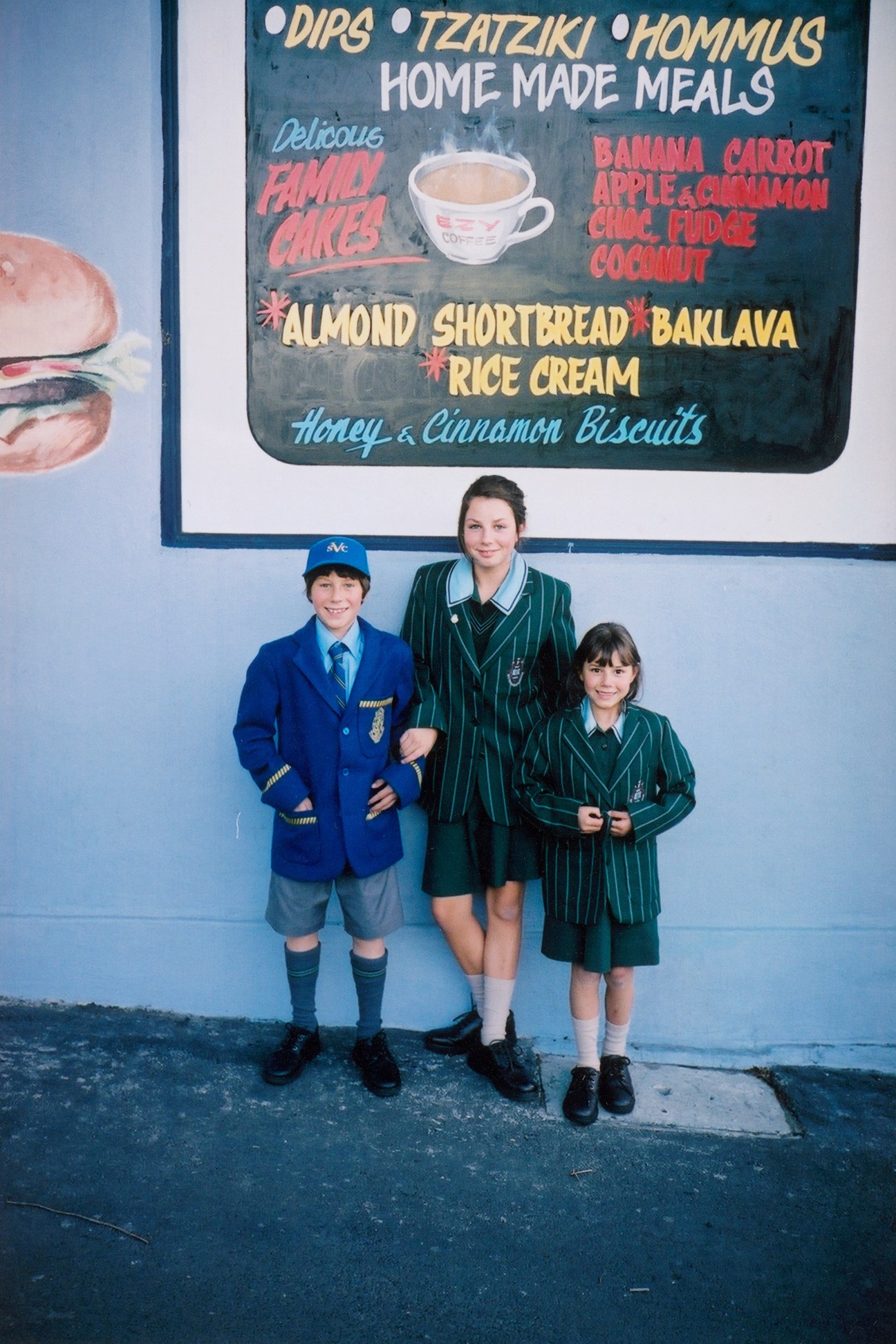 Three school children in uniforms stand outside a Greek cornershop with a blue menu board.