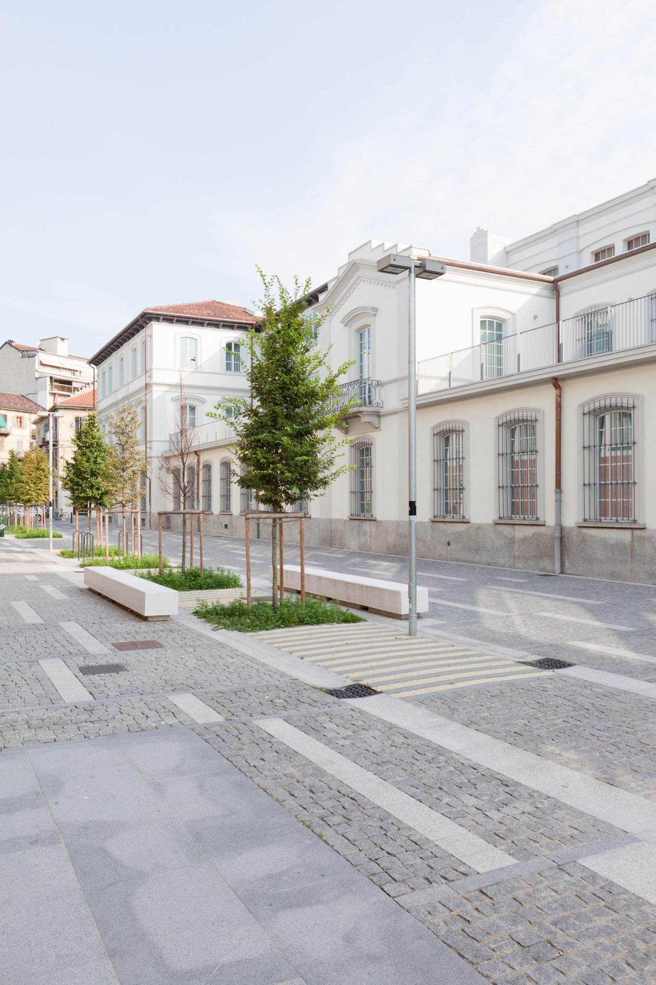 A modern public plaza with paved walkways, young trees in planters, and neoclassical white buildings with barred windows in the background.