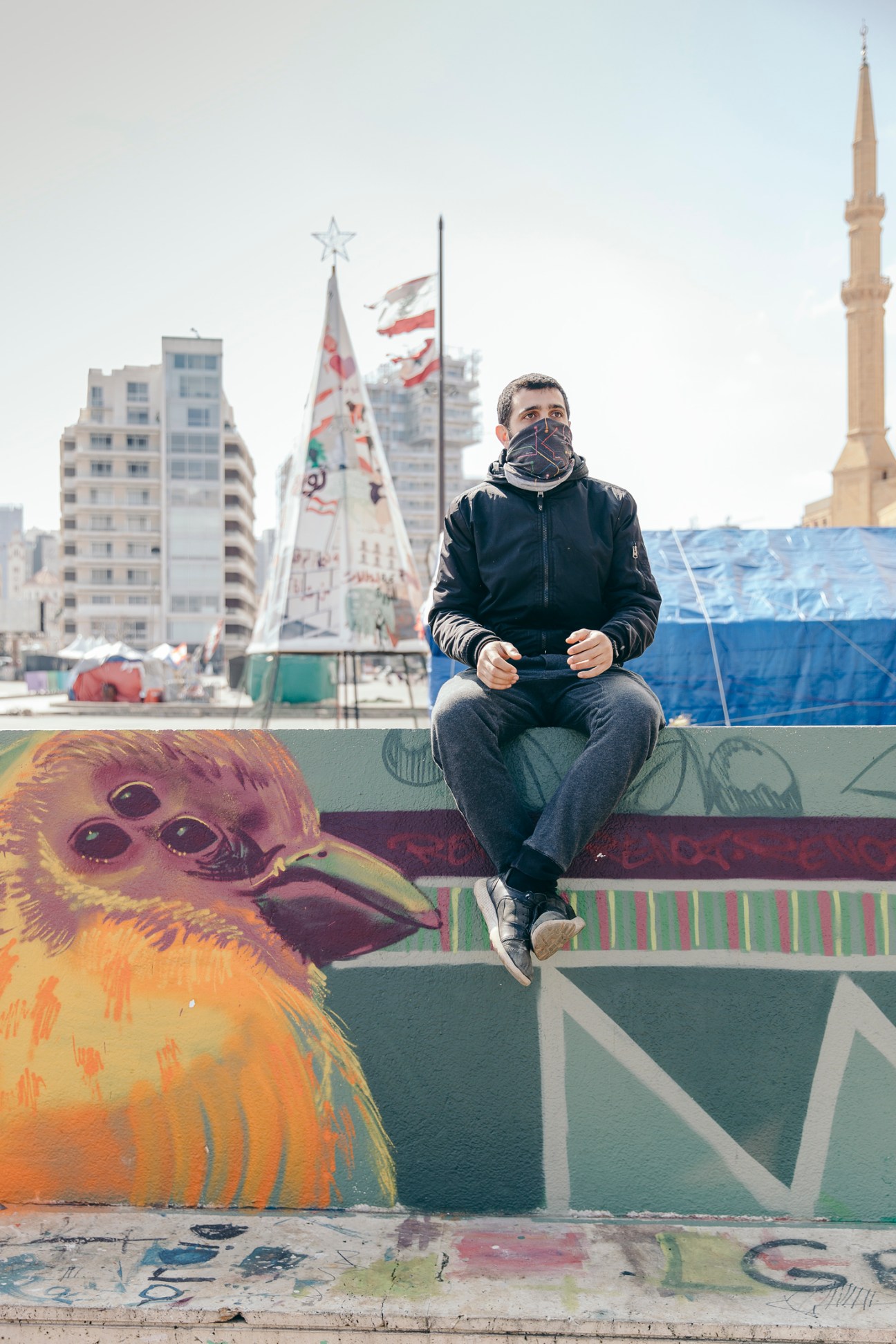 Ahmad Ghaddar sits on a colorful wall with street art including a painted bird character in Beirut