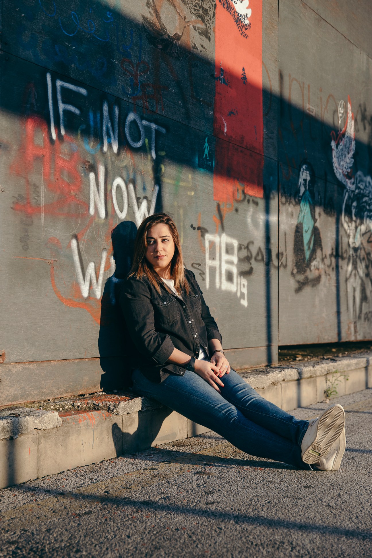 Rachel Karam sits against a graffitied wall in Beirut wearing denim jacket and jeans