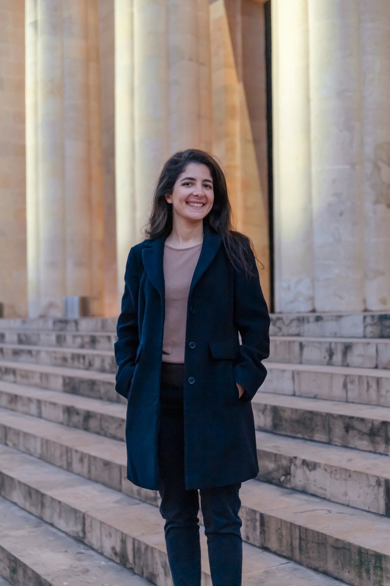 Nermine Sibai stands smiling on stone steps in front of classical columns wearing a dark coat
